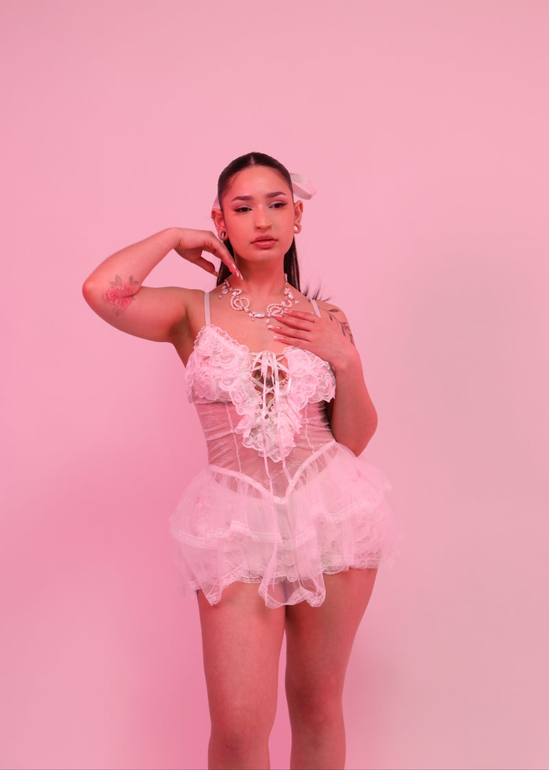A woman modeling a white lace lingerie set consisting of a lace corset top with a lace tutu skirt, posed against a solid pink background.